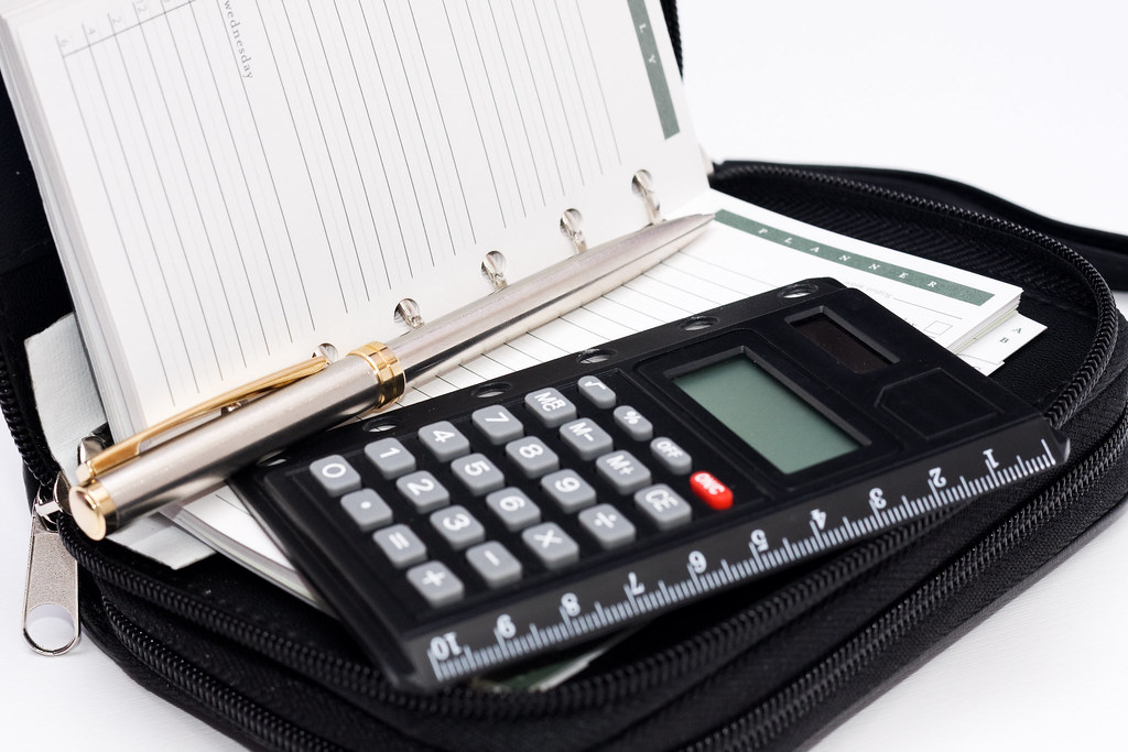 An open daily planner notebook, silver pen, and small electronic calculator placed on a black zipper portfolio organizer